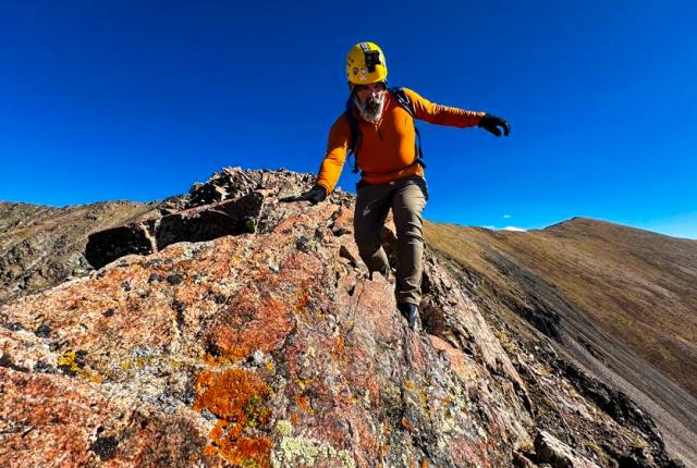 A climber in an orange shirt and yellow helmet ascends a rocky ridge, balancing carefully. The sky is clear blue, and the terrain is rugged and uneven.