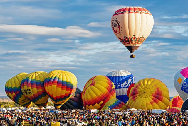 A crowd gathers to watch colorful hot-air balloons inflate and take flight against a blue sky.