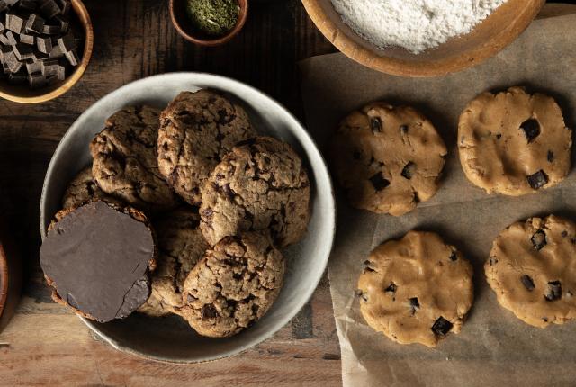 A bowl of chocolate chip cookies next to raw cookie dough on parchment paper. Nearby are bowls of flour, chocolate chunks, and herbs on a wooden table.