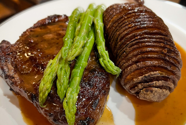 Grilled steak with sauce is topped with bright green asparagus spears, accompanied by a sliced, baked potato, all on a white plate.