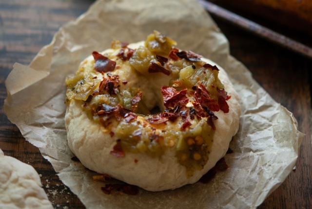 A bagel topped with chopped green chiles and red pepper flakes rests on brown parchment paper.