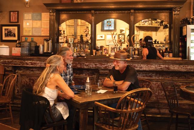 Three people sit at a wooden table in a dimly-lit, old-timey saloon.