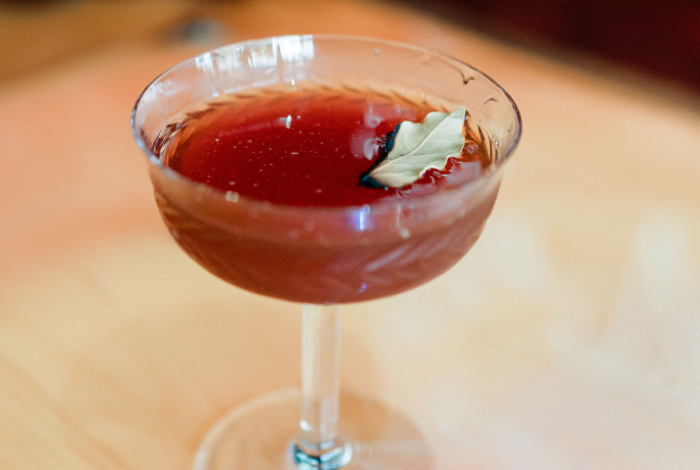 Amber cocktail garnished with a bay leaf in a patterned stemmed glass on a wooden bar.