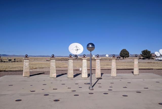 A metal sphere atop a slender post casts a shadow across a concrete surface dotted with markers.