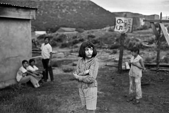 Black and white photograph of a young girl standing with folded arms in the foreground beside a dirt road in Petaca, New Mexico.