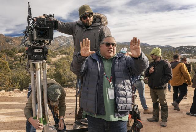 A film crew member in a blue puffer jacket holds up both hands in a 'stop' gesture while standing in front of a camera on a dirt road.