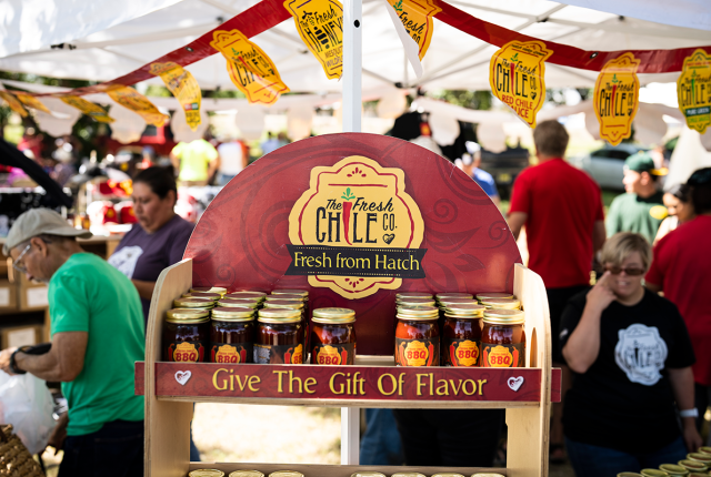Farmers’ market booth displaying jars of chile products from The Fresh Chile Co., with shoppers in the background.