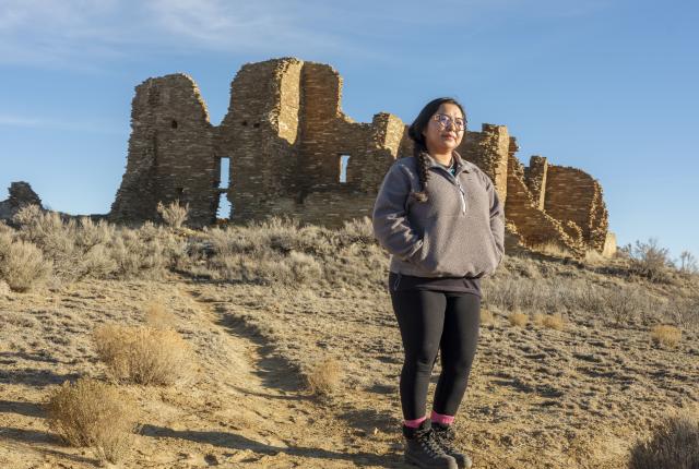 A person stands on a sandy, shrub-filled landscape before ancient stone ruins under a clear blue sky.