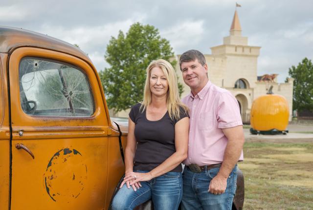 A man and a woman stand in front of an orange vintage truck, with a large, sculpted pumpkin and a castle-like building in the background.
