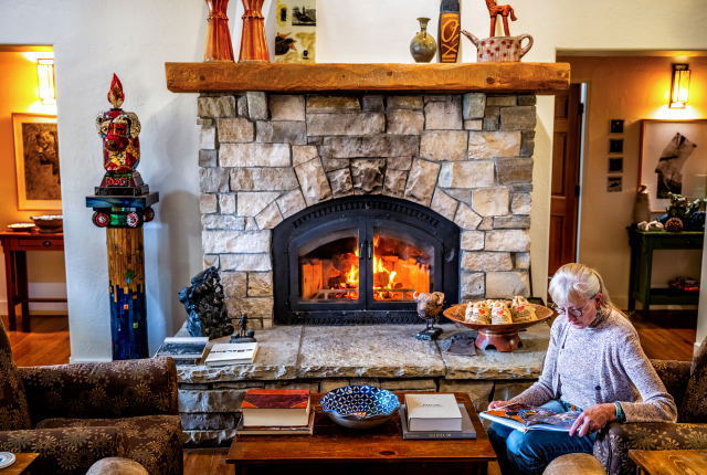 Cozy living room with a warm stone fireplace, books, and decorative items. A person sits reading on a chair.