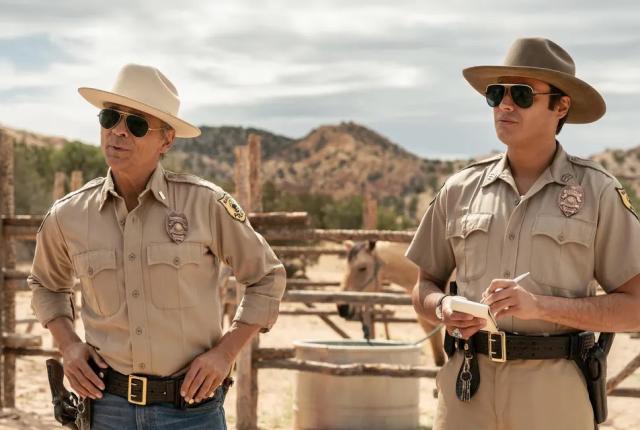 Two men in sheriff uniforms and sunglasses stand outside in a sunlit desert setting, with a horse in the background.