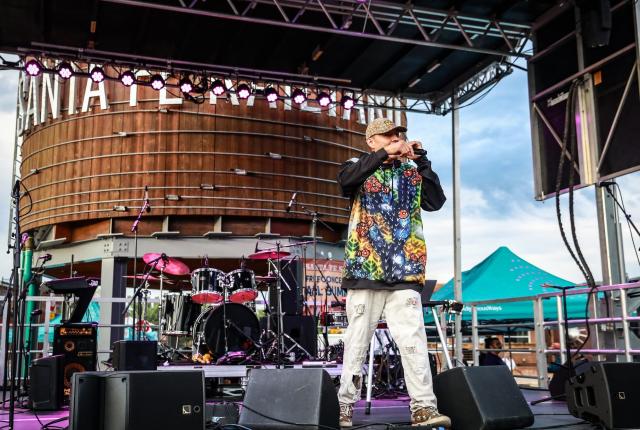 Performer on an outdoor stage with instruments and lighting, standing before a large wooden structure marked ‘Santa Fe.’