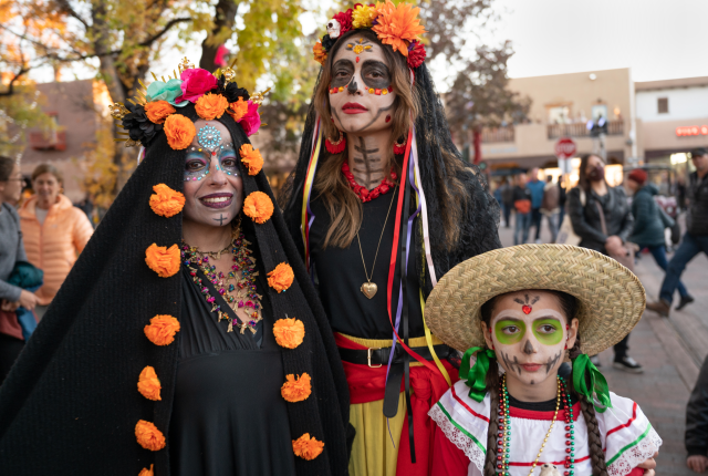 Three people, two women and a girl, pose in traditional Dia de los Muertos attire and makeup.