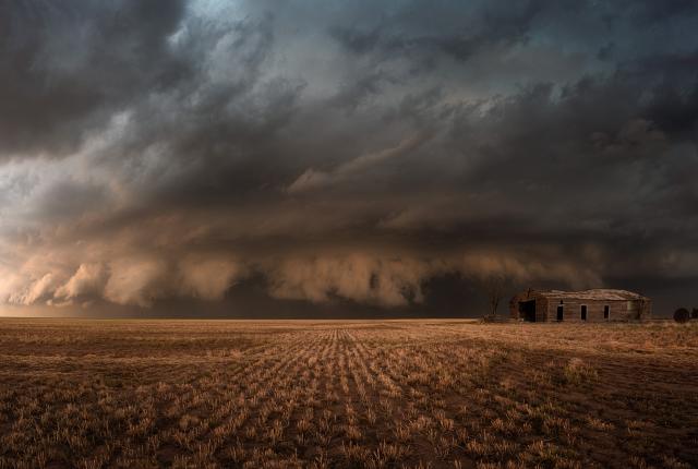 A massive, dark storm cloud hovers over an abandoned farmhouse and a barren field.