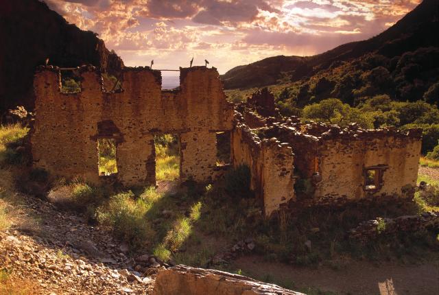 The sunlit Van Patten Ruins in a green valley, silhouetted against a dramatic sunset sky.