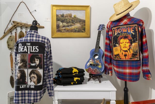 Blue acoustic guitar decorated with bright floral designs next to a red flannel shirt embroidered with a David Bowie design on the back.