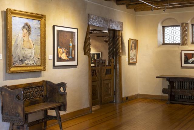 A room with wood floors and white walls containing framed paintings and rustic wooden furniture.