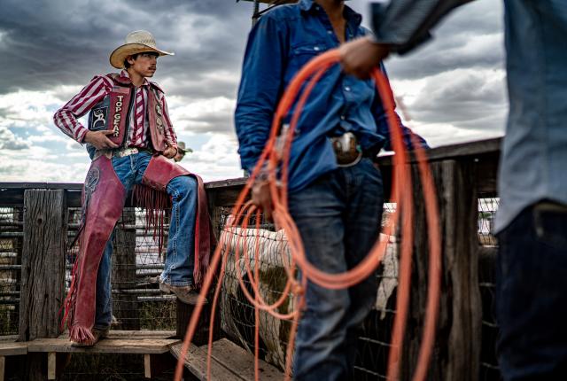 Rodeo participants stand by a wooden fence as one person in red chaps waits with others holding coiled ropes under a cloudy sky.