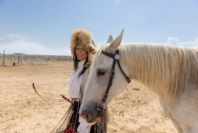 Person standing beside a light‑colored horse in a dry, open landscape, holding a bow with arrows and wearing a fur hat and decorated clothing.