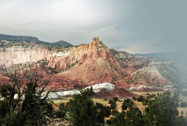 Multicolored desert cliffs and buttes rise above rolling red, white, and tan hills dotted with shrubs under an overcast sky.