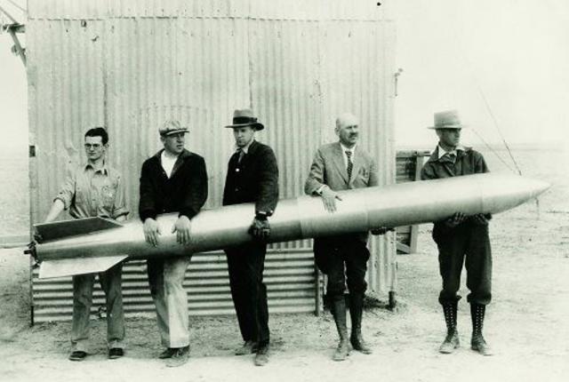 Five men hold a large, cylindrical rocket horizontally in front of a corrugated metal building.