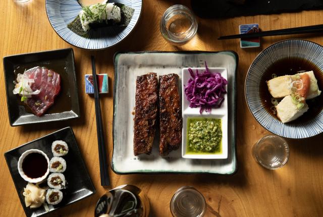 Tabletop spread of assorted dishes including grilled ribs, sliced fish, sushi rolls, and sides.