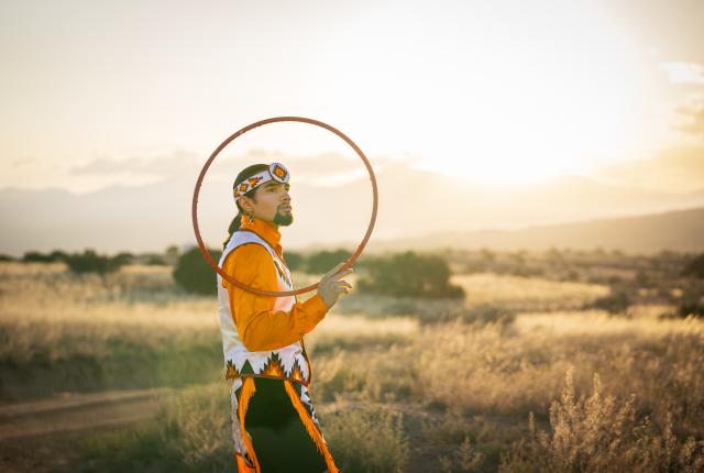 erson outdoors at sunset holding a hoop, wearing a brightly colored outfit with detailed beadwork and patterned accessories, standing in a golden field with mountains softly visible in the background.