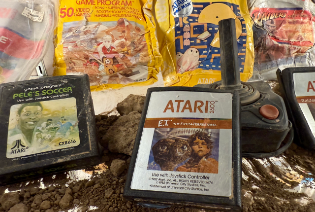 A collection of vintage Atari 2600 game cartridges lies on the ground, partially buried in dirt. Visible titles include "Pele's Soccer" and "E.T. the Extra-Terrestrial." A nearby joystick is also visible.