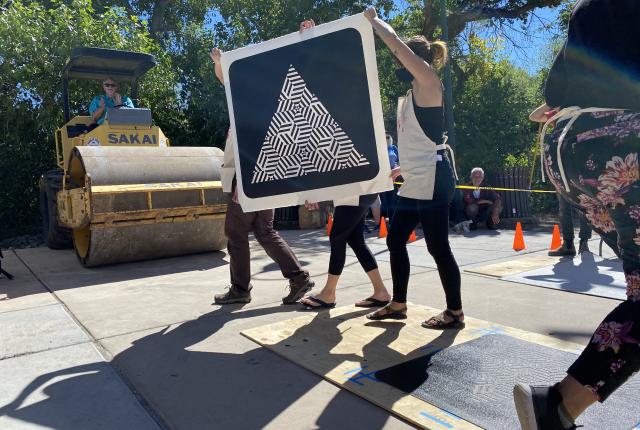 People carry a large black-and-white geometric print past a steamroller outdoors.