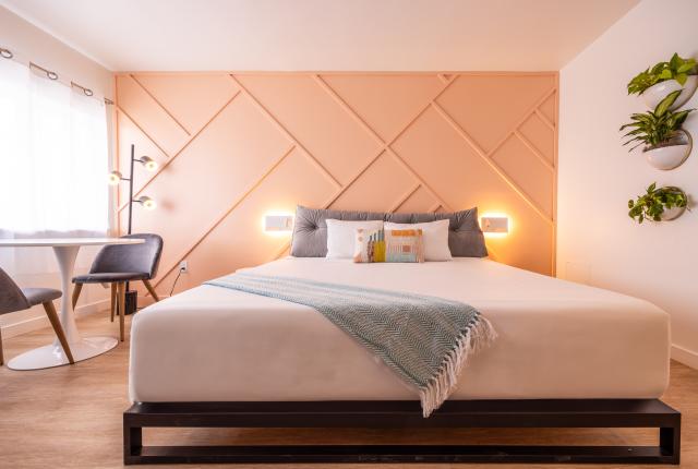 A vibrant, open guest room with minimalist retro styling, including a bedside reading nook, colorful geometric accents, and simple modern furnishings.