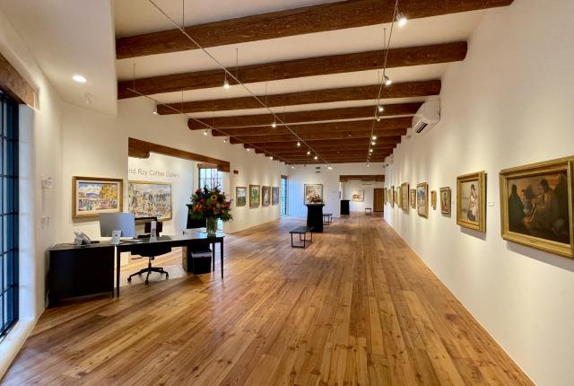 Bright, wood‑floored gallery with paintings lining both walls and exposed ceiling beams above.