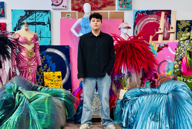 A person in a black shirt and jeans stands in an art studio surrounded by vibrant, textured fabrics and abstract art.