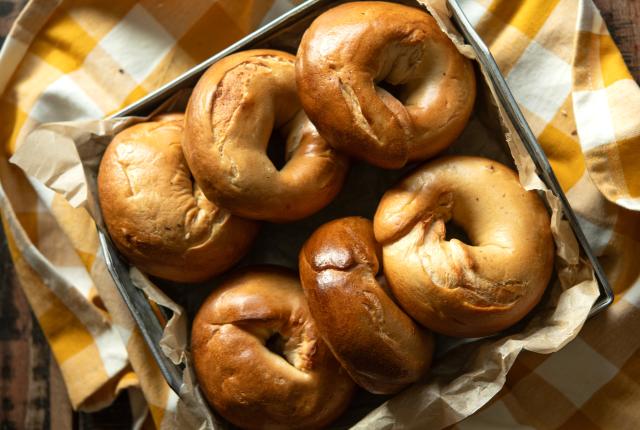 A metal tray of six fresh bagels sits on a yellow gingham cloth.