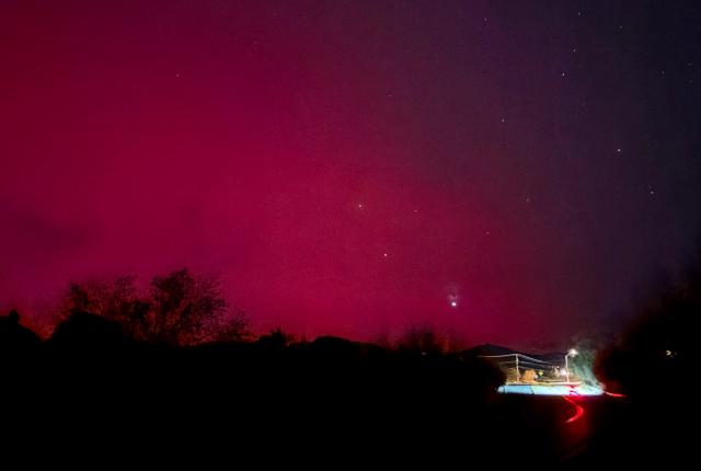 A vivid pink night sky with scattered stars illuminates a rural area. Silhouetted trees line the horizon, and a vehicle's red taillights trace a road.