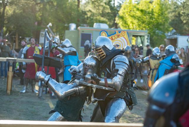 Two armored knights engage in intense combat at an outdoor medieval festival, with onlookers watching and a colorful food truck in the background.