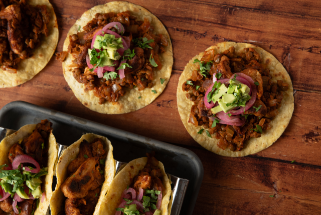 Close-up of three tacos filled with BBQ lion's mane mushrooms and lentils, topped with pickled onions, avocado, and cilantro on a wooden table.