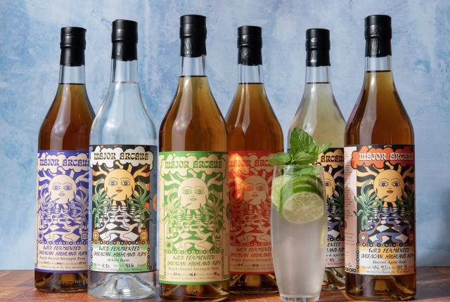Five bottles with colorful labels stand against a textured blue background. A clear liquid is being poured into a glass with lime and mint.