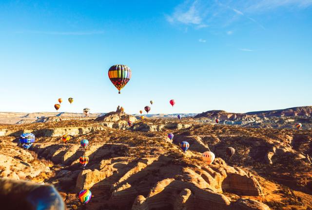 Colorful hot air balloons float above a sunlit desert landscape with rocky formations and clear blue sky.
