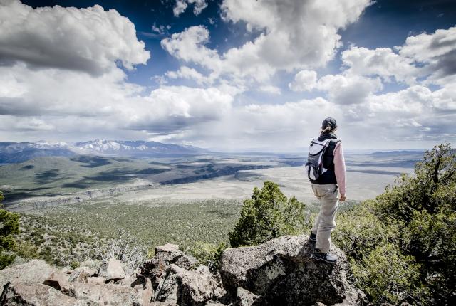 Hiker with a backpack stands on rocky terrain overlooking a wide valley, distant mountains, and dramatic cloud-filled sky.