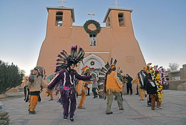 A group of Indigenous dancers wearing colorful feathered headdresses and traditional attire perform in front of a historic adobe church with wreaths, beneath a clear sky.