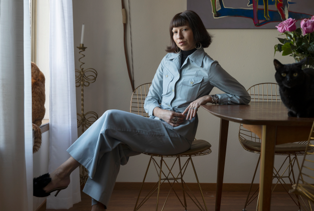 A woman in a light blue jumpsuit sits at a wooden table, with a calm black cat beside her.