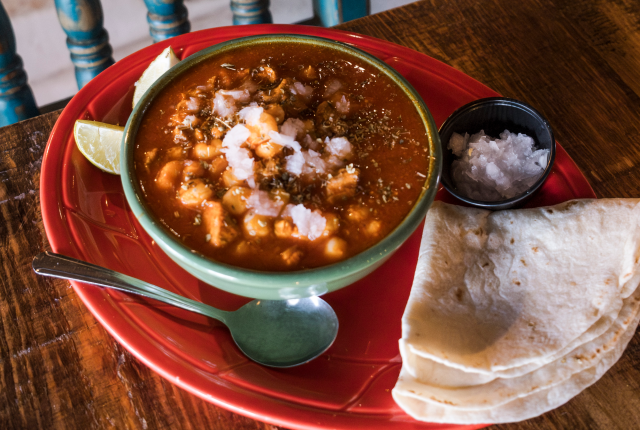 A bowl of red posole sits on a red plate with a side of corn tortillas and chopped white onion.