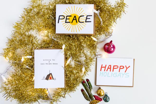 Three holiday cards with the sayings "Peace," "Wishing you a cozy holiday season," and "Happy Holigays" are arranged on a white surface with gold tinsel, string lights, a metal cactus, and vintage ornaments.