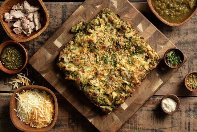 Square green chile pizza on a wooden board surrounded by bowls of cheese, chicken, pesto, chiles, and herbs.