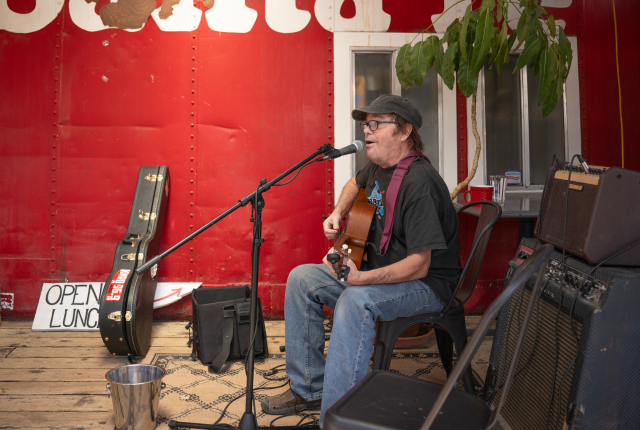 A man sits on a chair playing an acoustic guitar in front of a red wall with "Santa Fe" painted on it.
