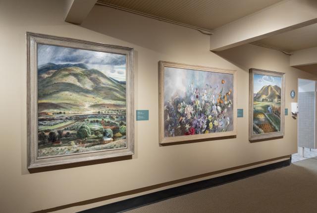 Hallway gallery displaying three framed paintings: two landscape scenes with rolling hills and one large floral painting.