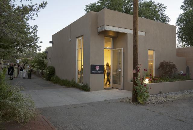 A modern adobe building labeled “smoke the moon” glows with interior light at dusk, with a group of people gathered along the driveway outside.