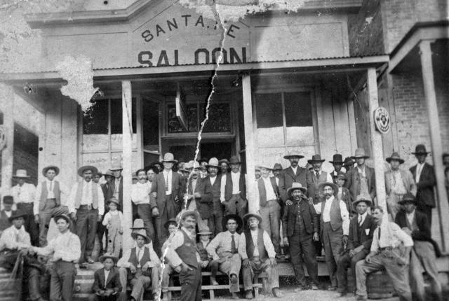 A black and white photo of a large group of men in hats, most with mustaches, posed in front of the Santa Fe Saloon.