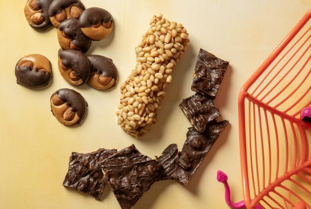 Assorted chocolate-dipped candies, a nut-covered candy roll, and pieces of chocolate bark on a pale yellow surface.