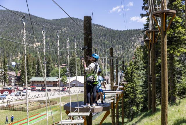 Two people in helmets and harnesses navigate a high ropes course against a backdrop of pine-covered mountains in Red River.